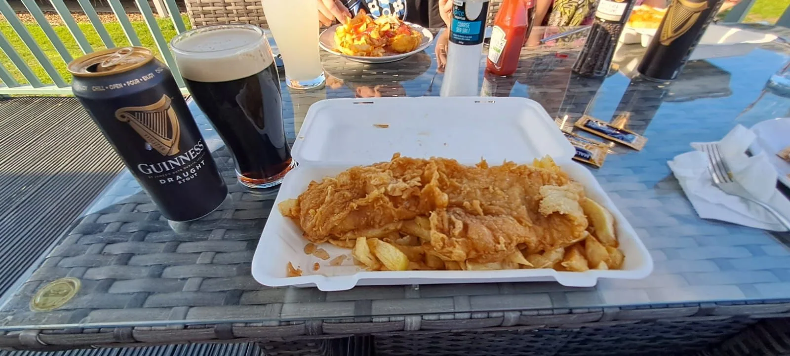 Fish and chips in a takeaway tray with a pint on a glass-topped outdoor table