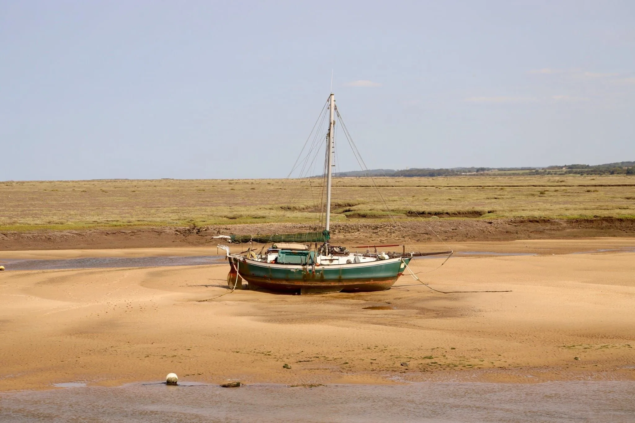 An old green-hulled sailing boat resting on the sand at low tide, with salt marsh and open sky behind