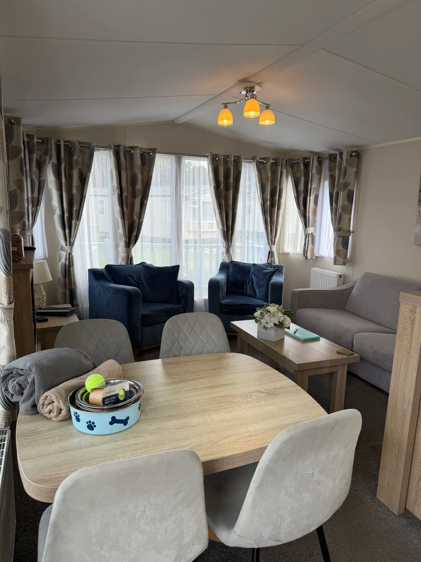Open-plan living and dining area inside Tern 32 with a wooden table for six, grey chairs, a grey sofa, two deep blue armchairs, bone-print dog bowls and patterned curtains