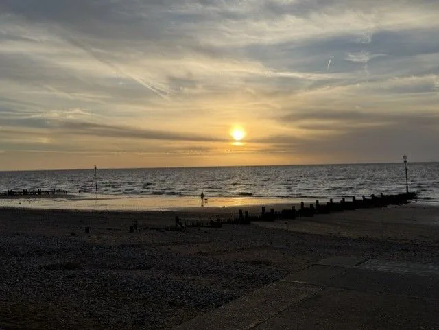 Sunset over the sea from a beach, with wooden sea defences, wet sand and a distant walker