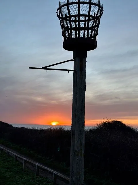 The silhouette of a basket-topped wooden post on the clifftop against a pink and orange sunset sky