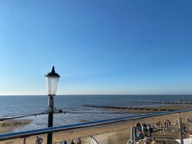A Victorian-style lamppost overlooking people on the beach and promenade, with wooden sea defences and calm sea beneath a clear blue sky