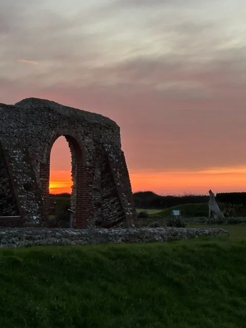 Old stone and brick ruins with a tall arch standing in grass against an orange sunset