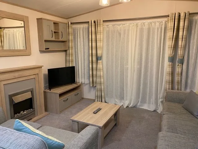 The lounge inside Windsor 30 with an electric fire in a wooden surround, a wall mirror, a television on a wooden unit, a coffee table and grey sofas